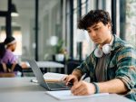 Jeune étudiant prenant des notes tout en apprenant en ligne sur un ordinateur portable à l’université.