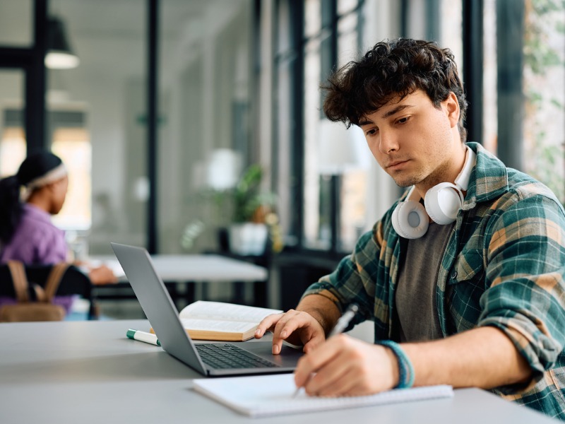 Jeune étudiant prenant des notes tout en apprenant en ligne sur un ordinateur portable à l’université.