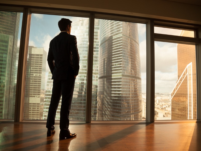 Homme d’affaires pensif regarde par la grande fenêtre à la ville de coucher de soleil.