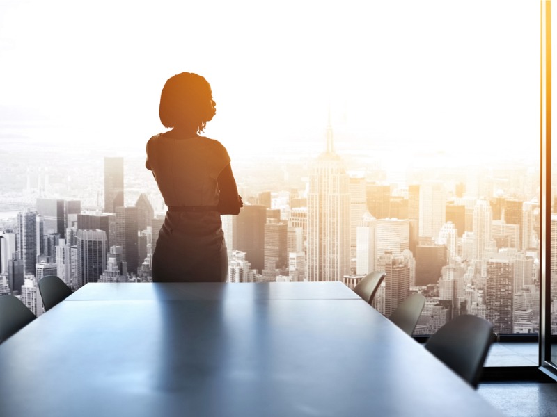 Une femme d'affaire devant une fenêtre qui regarde la ville depuis un bureau dans un building.