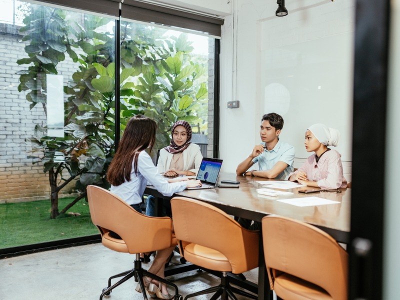 Un groupe de personnes diverses s’est engagé dans une réunion d’affaires ciblée dans un bureau moderne avec de grandes fenêtres et des plantes vertes à l’extérieur.